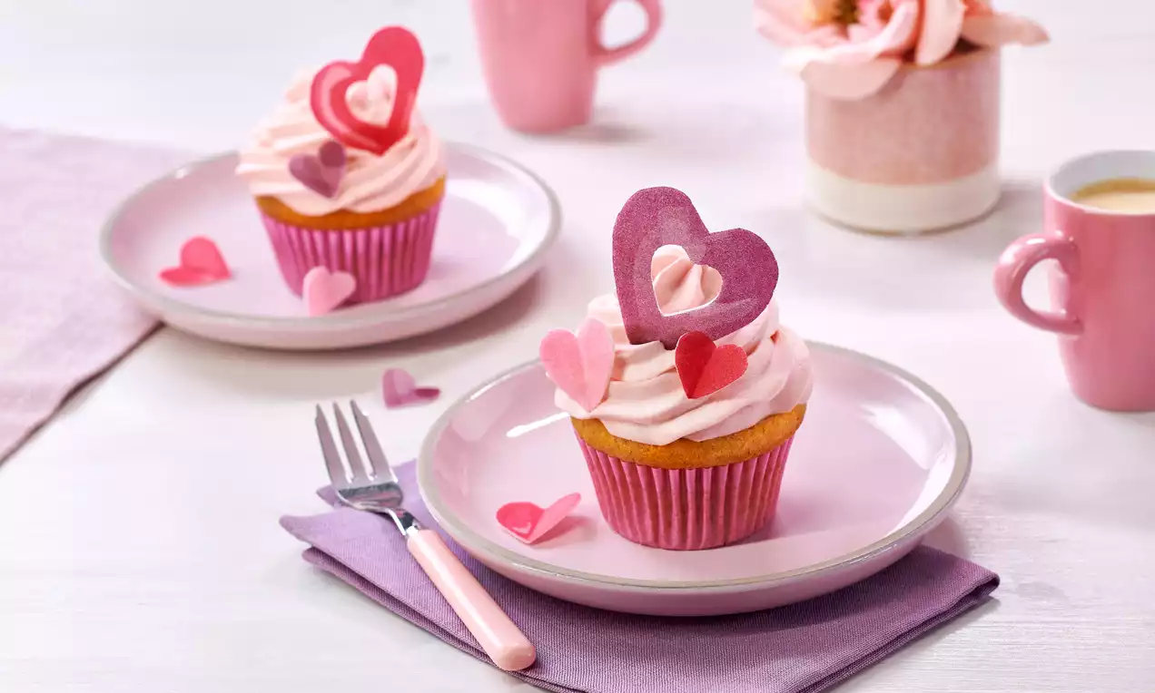 Ein Cupcake mit zartrosa Cremehaube, verziert mit rosa und roten Zuckerherzen, serviert auf einem rosa Teller mit violetter Serviette