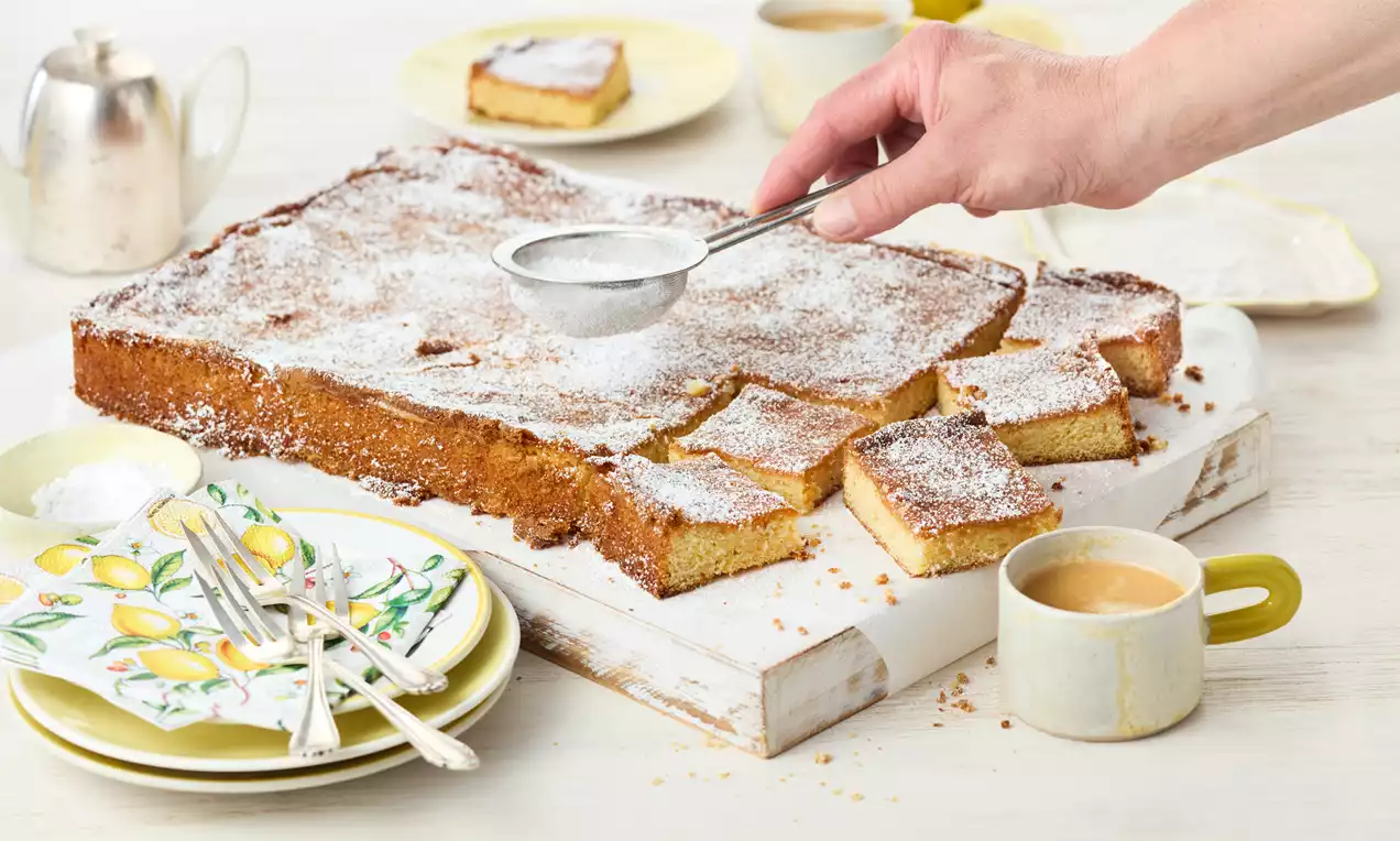 Zitronen-Brownie mit heller, saftiger Krume, weißen Schokoladenstücken, Zitronenzeste und Puderzucker auf weißem Teller