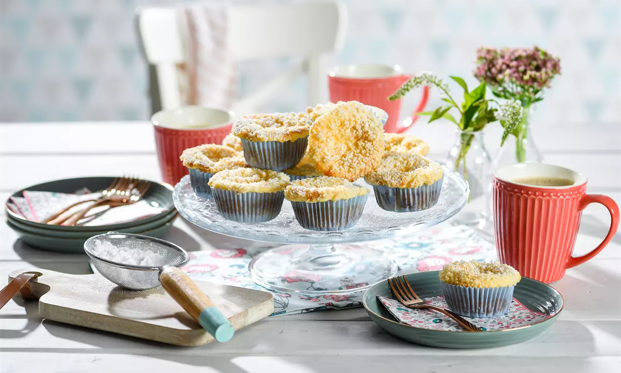 Käsekuchen-Muffins mit goldgelber Streuseldecke in blauen Papierförmchen, auf einer Glasplatte mit Tassen und Tellern im floralen Design drapiert
