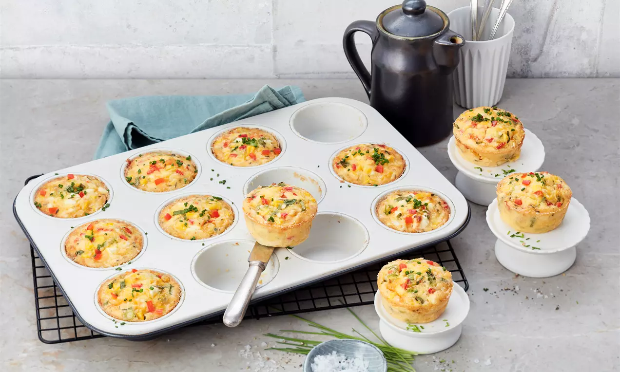 Goldene, fluffige Frühstücks-Muffins mit Kräutern und Gemüse in einer weißen Backform, serviert auf kleinen Tellern neben einer Kanne