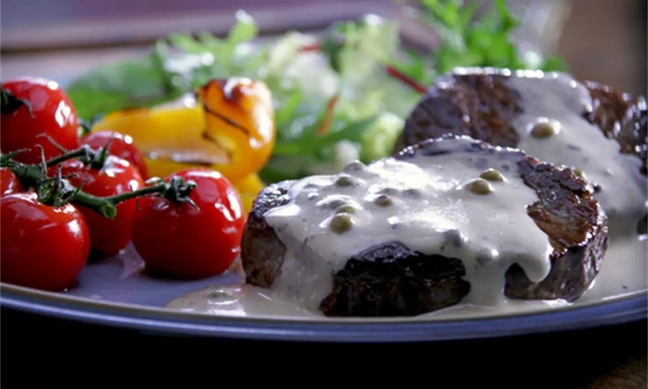 Zwei dunkelbraun gebratene Rindersteaks mit sahniger Pfeffersoße, frische Kirschtomaten, Grillpaprika und bunte Salatblätter auf grauem Teller