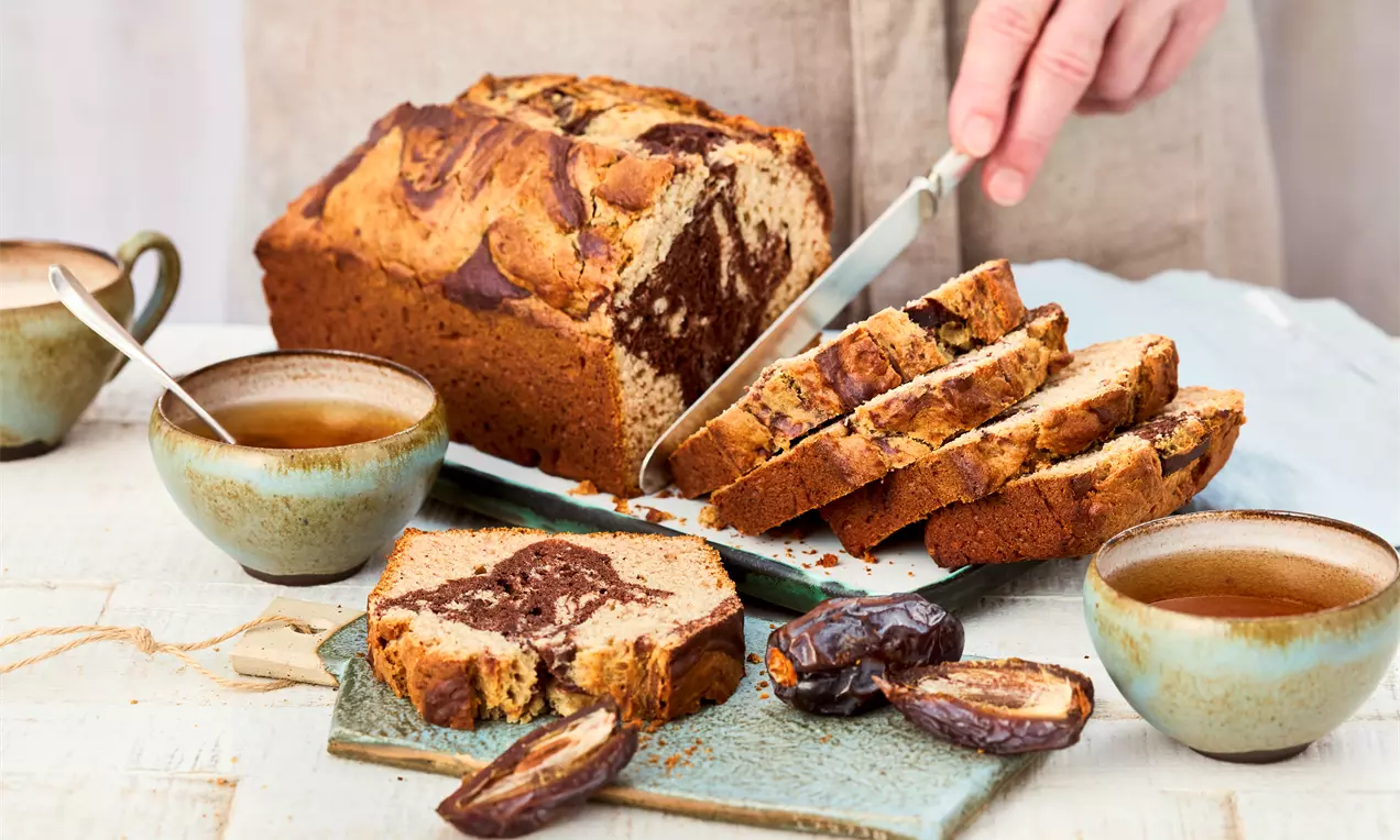 Kuchen ohne Zucker auf einem Brett, angeschnitten und in Scheiben. Marmormuster im Teig sichtbar. Daneben Datteln, zwei Tassen Tee und ein Messer. Eine Hand schneidet den Kuchen.