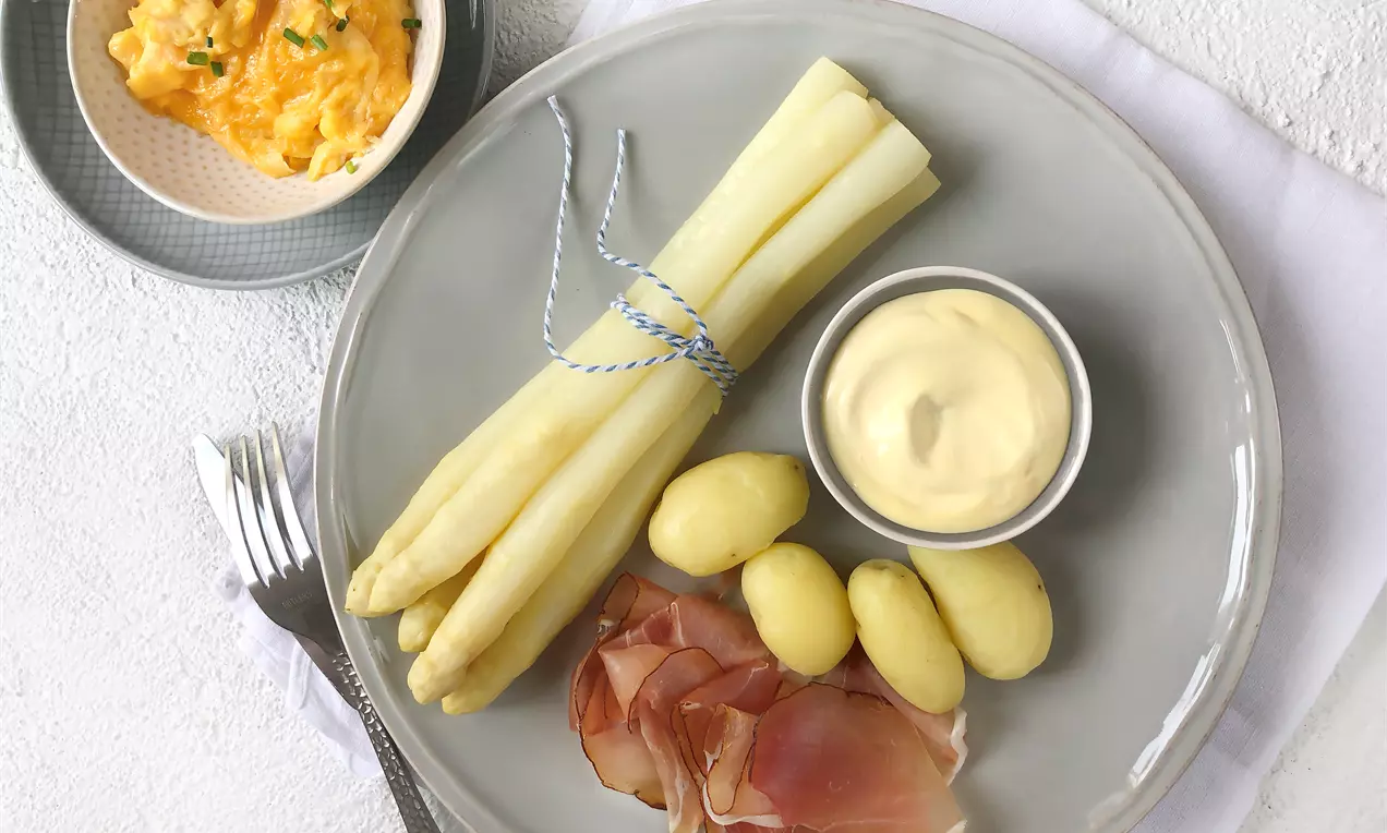Weißer Spargel mit blauem Band gebunden, hellgelbe Kartoffeln, rosa Schinken und cremige Sauce auf grauem Teller mit weißem Stoffunterlage