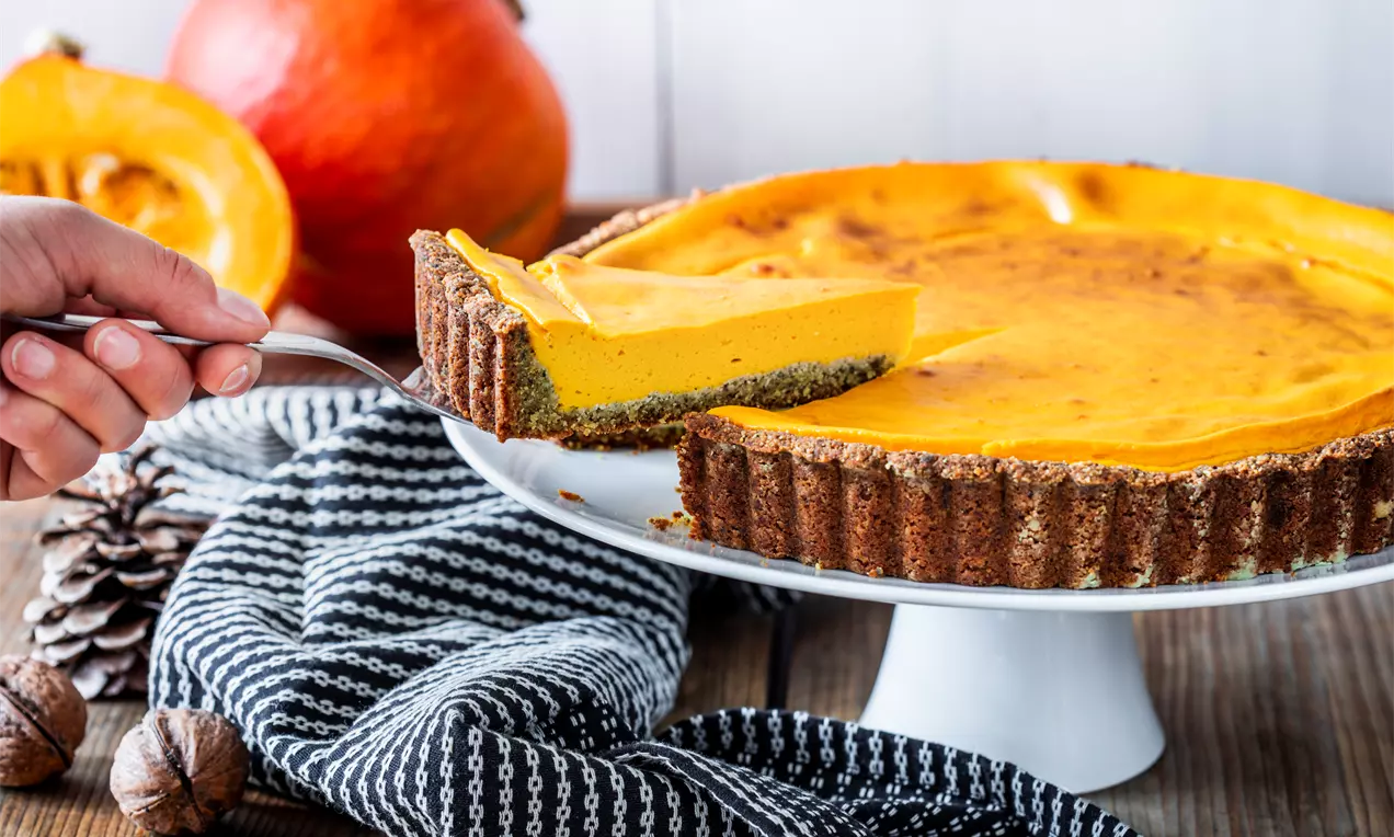 Glutenfreie Kürbis-Tarte mit leuchtend orangefarbener Füllung und braunem Nussboden auf weißer Tortenplatte, dekoriert mit Kürbis und Tuch