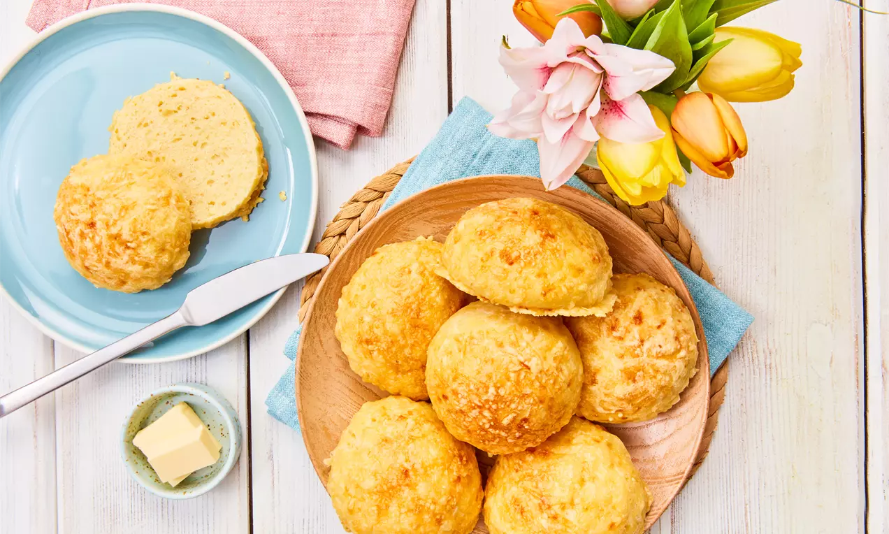 Käsebrötchen mit goldgelber Kruste auf einem runden Holzbrett, daneben ein Teller mit halbiertem Brötchen, Butter und frischen Tulpen