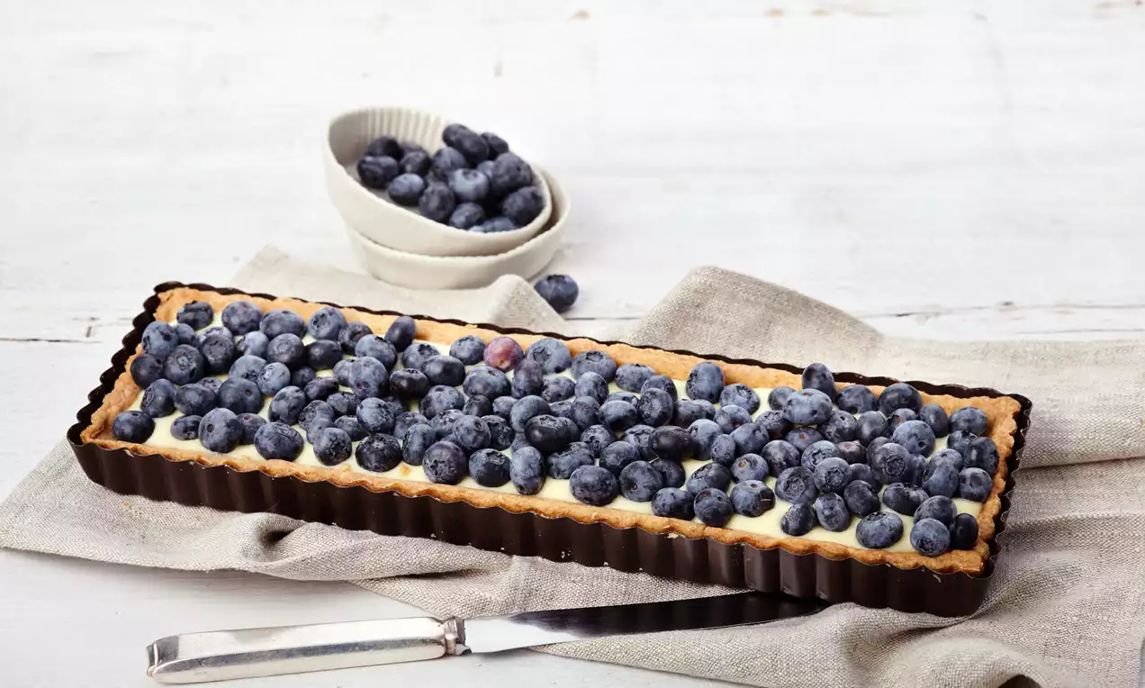 Rechteckige Tarte mit goldbraunem Mürbeteigboden, cremiger Puddingfüllung und einer dichten Schicht frischer Blaubeeren auf Leinentuch drapiert