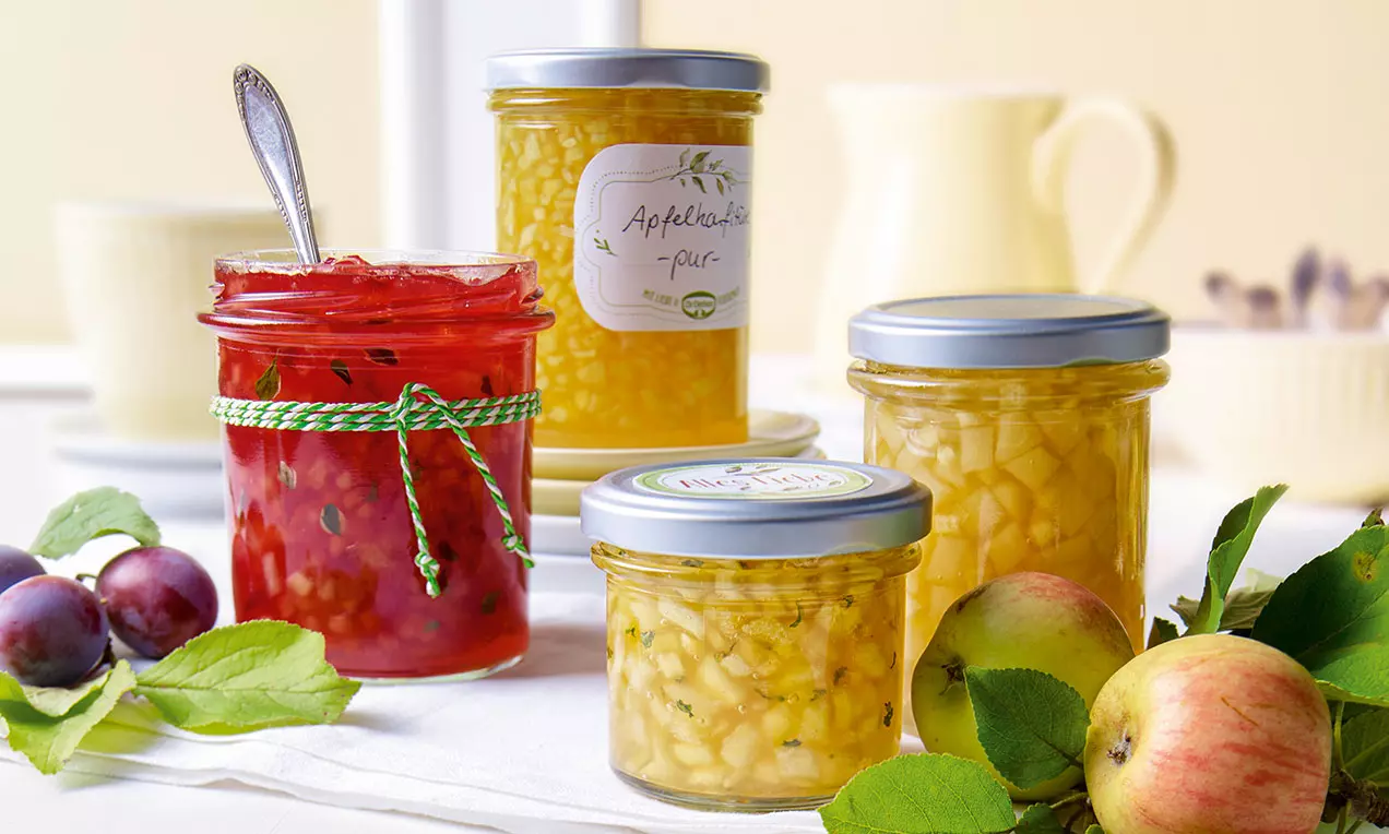 Glas mit roter Apfel-Konfitüre neben hellgelben Gläsern, umgeben von Äpfeln, Pflaumen und grünen Blättern auf einem weißen Tuch