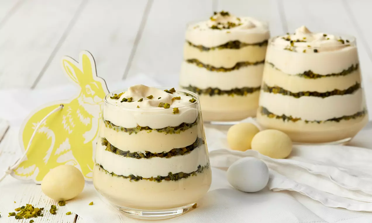 Eierlikör-Pudding in einem Glas schichtweise mit heller Creme und fein gehackten Pistazien dekoriert, mit Ostereiern auf weißer Serviette arrangiert