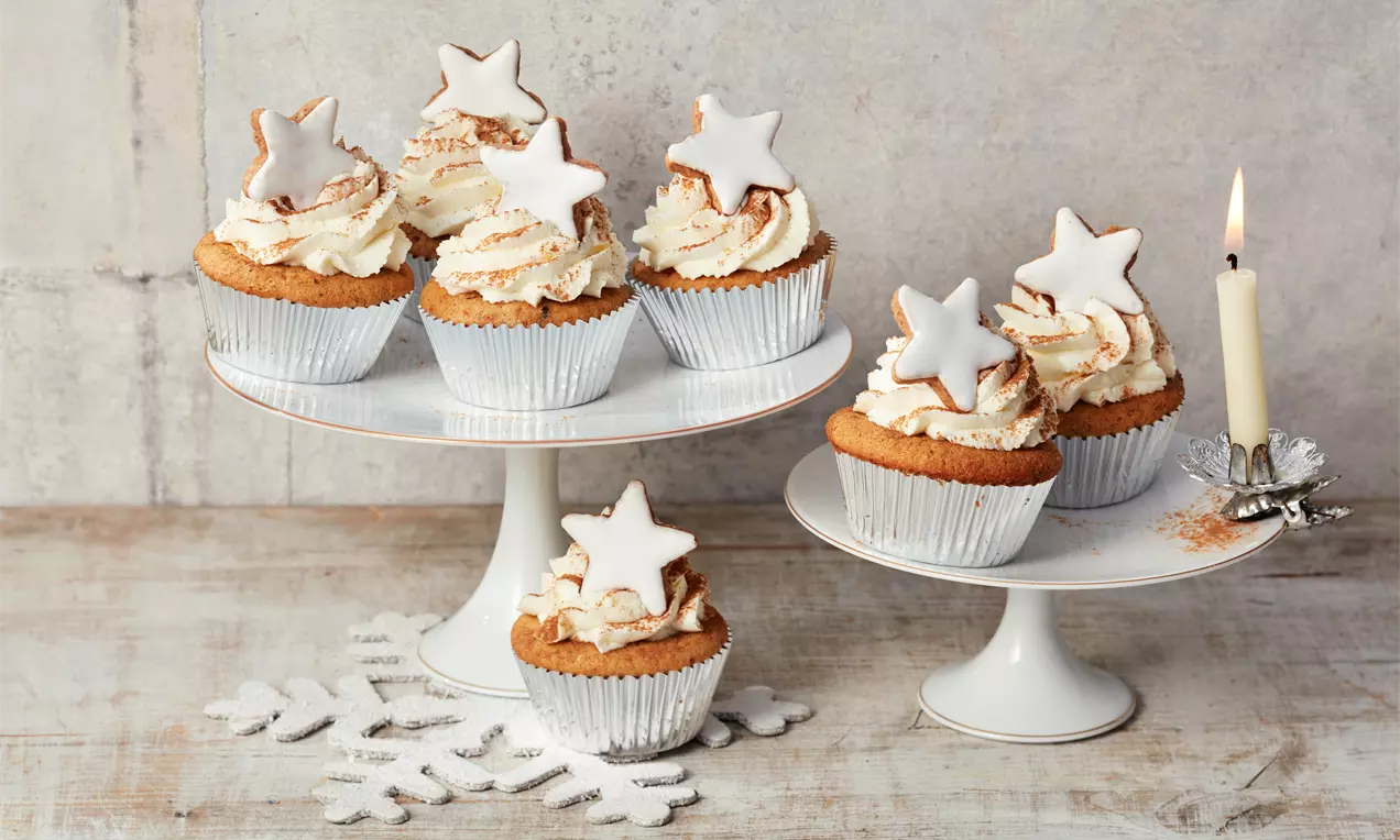 Weihnachtsmuffins mit leicht bräunlichem Teig, weißer Sahnehaube und Zimt, dekoriert mit einem weißen Zuckerstern auf weißen Tortenständern