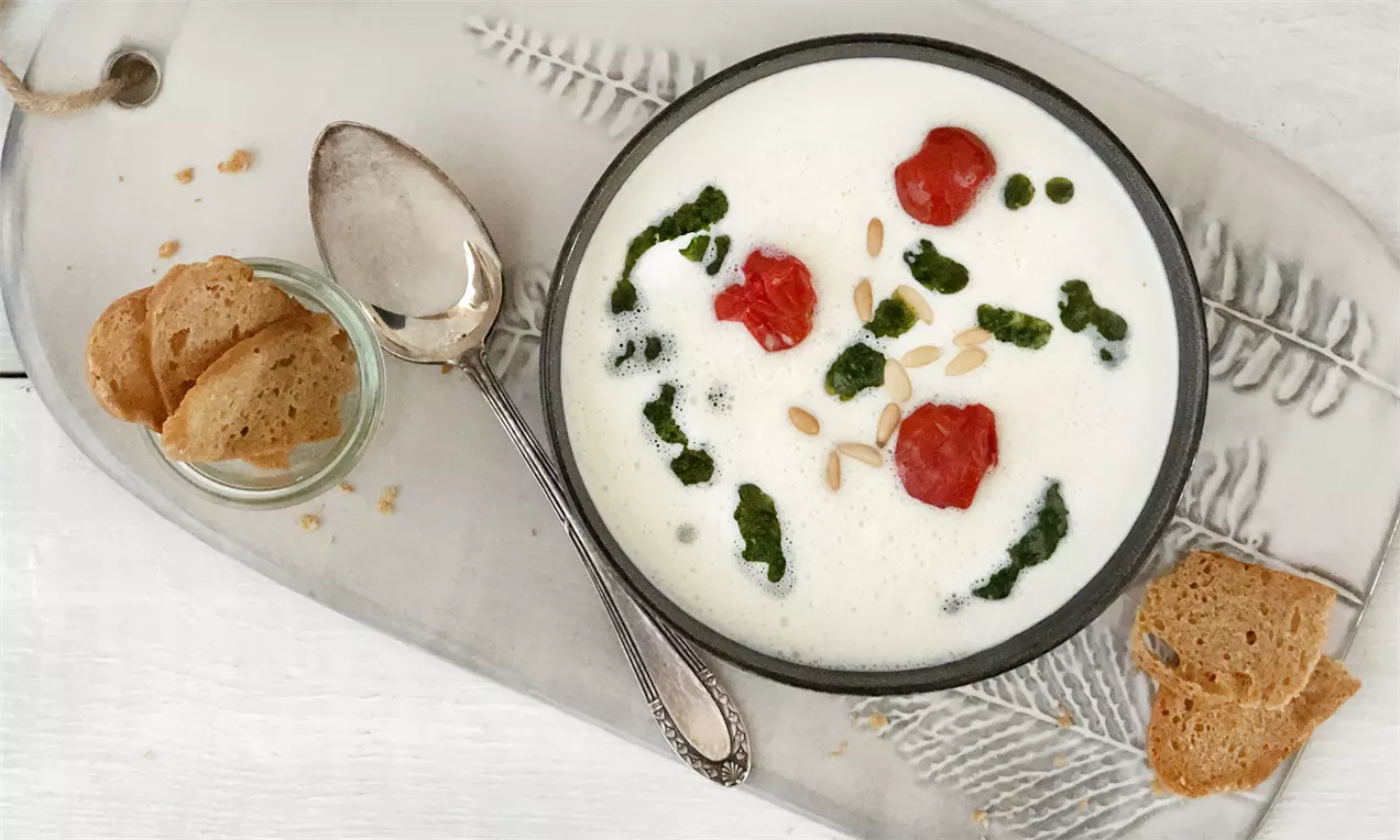 Cremig-weißes Mozzarella-Süppchen in schwarzer Schale mit roten Tomaten, grünem Pesto und Pinienkernen, serviert auf silbernem Tablett mit Brot