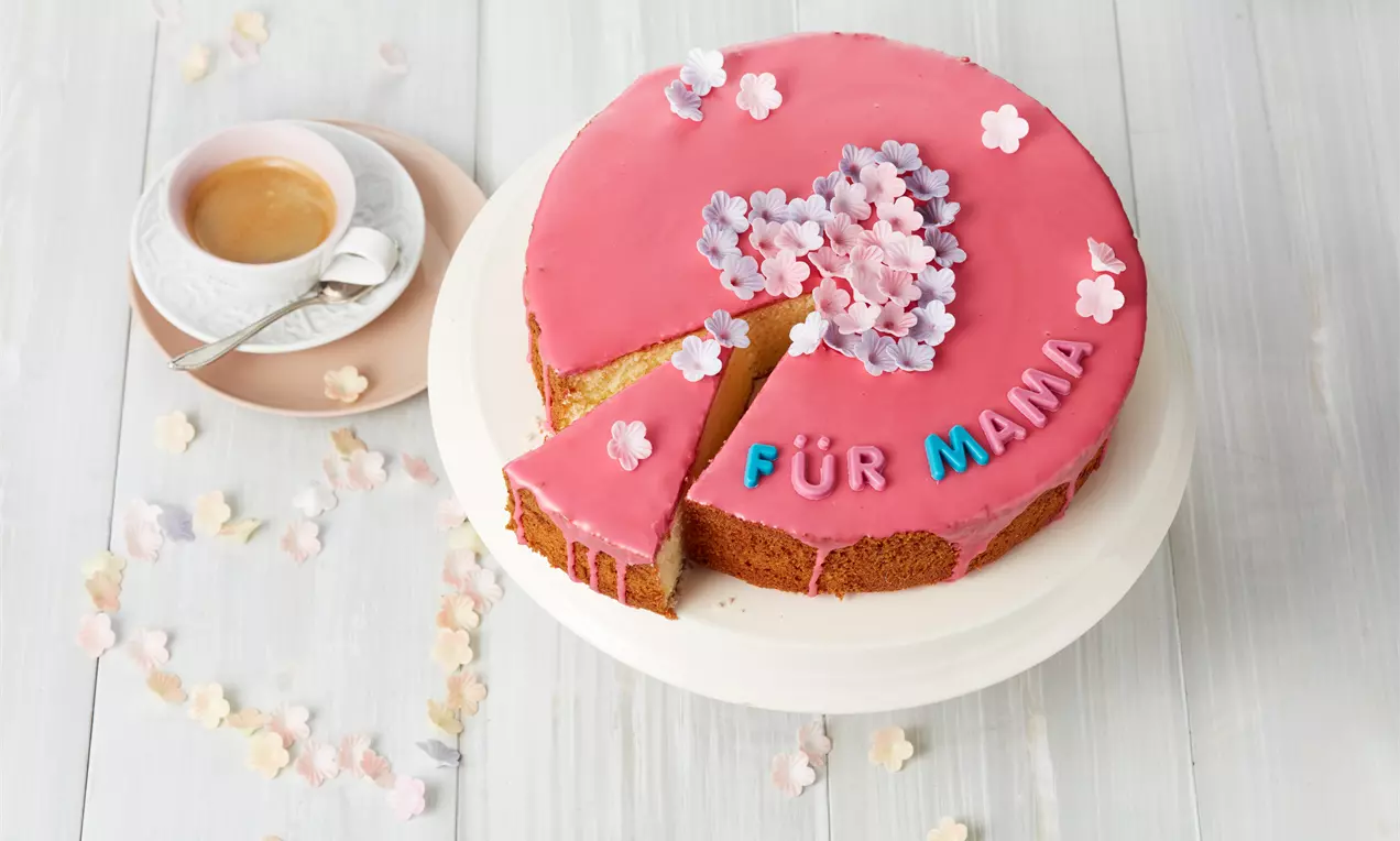 Ein runder Kuchen mit rosa Zuckerguss, bunten Buchstaben "Für Mama" und kleinen Zuckerblüten auf einer weißen Tortenplatte