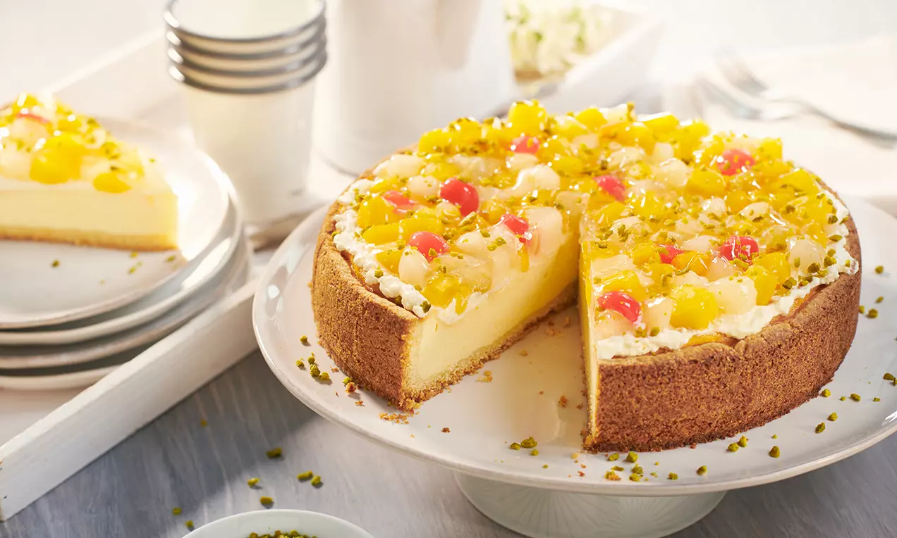 Käsekuchen mit goldbrauner Kekskruste, belegt mit gelben Fruchtstücken, roten Kirschen und Pistazien auf einer weißen Tortenplatte