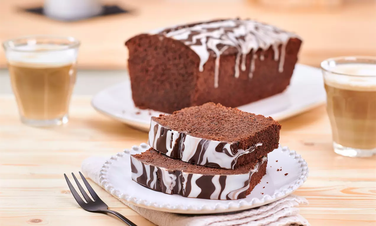 Saftiger, dunkelbrauner Schokokuchen mit marmorierter Glasur, in Scheiben geschnitten, auf einem weißen Teller mit Kaffeegläsern im Hintergrund
