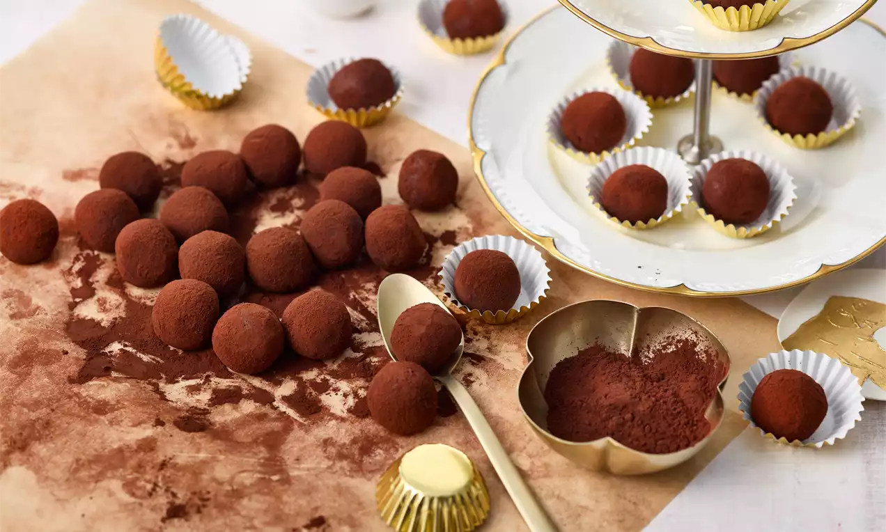 Kleine, runde Tiramisu-Kugeln mit feinem Kakaopulver bestäubt, auf gebräuntem Papier und in goldenen Förmchen neben einer Etagere angerichtet