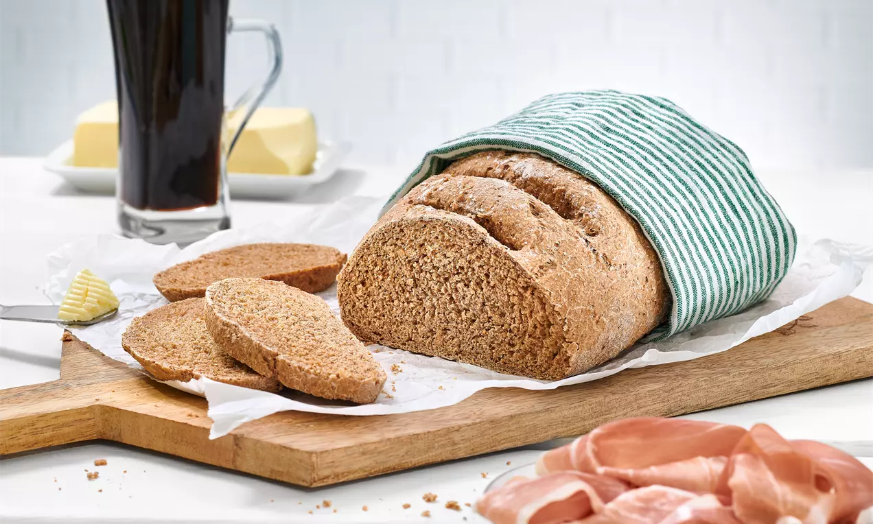 Ein ovales, goldbraunes Bierbrot mit weicher Krume in einem grün-weiß gestreiften Tuch auf einem Holzbrett, flankiert von Schinken und dunklem Bier