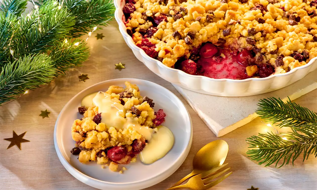 Goldbrauner Crumble mit roten Früchten in weißer Keramikform, portioniert auf Teller mit Vanillesoße, dekoriert mit Tannenzweigen und Sternen