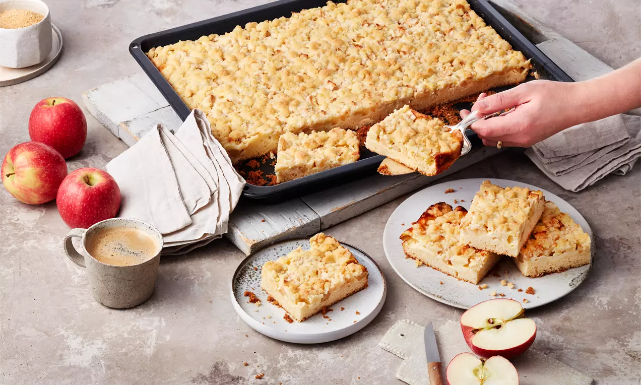 Goldbrauner Apfel-Streuselkuchen mit lockerer Schmandfüllung auf rechteckigem Backblech, serviert auf Tellern neben roten Äpfeln und Kaffee