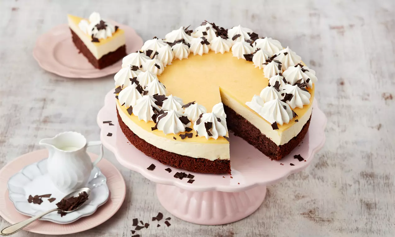 Eierlikörtorte mit gelber Creme, weißer Sahnehaube und Schokoraspeln auf dunklem Boden auf rosa Tortenplatte mit Teekanne und Teller