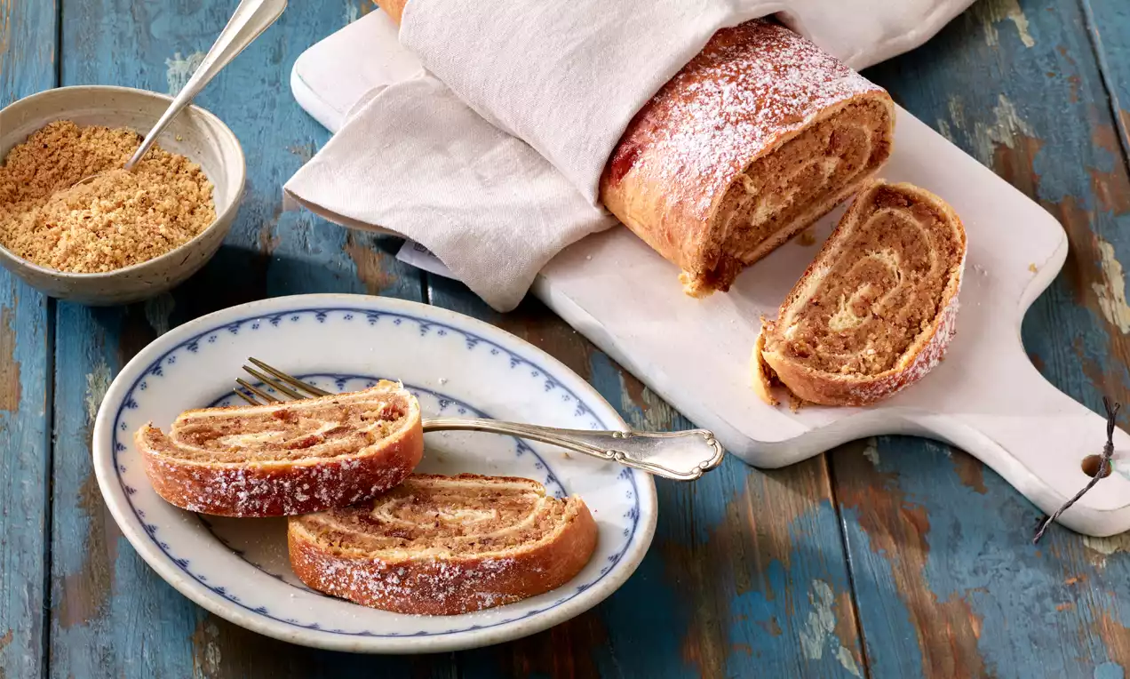 Goldbrauner Nussstrudel mit zarten Spiralmustern, bestäubt mit Puderzucker, auf Holzbrett und Teller mit Besteck angerichtet