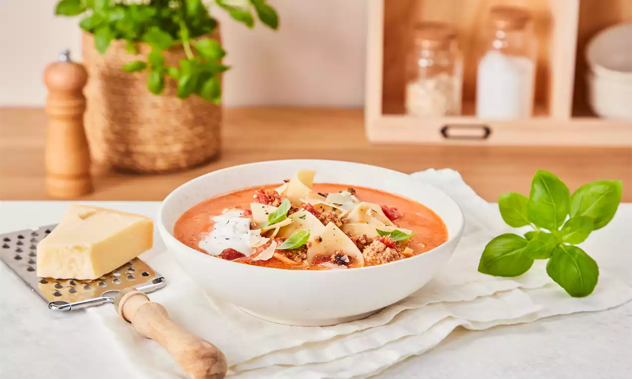 Lasagnesuppe mit roter Tomatencreme, Nudestücken, Hackfleisch, Kräutern, Parmesanraspeln und Sahnehäubchen in einer weißen Schale
