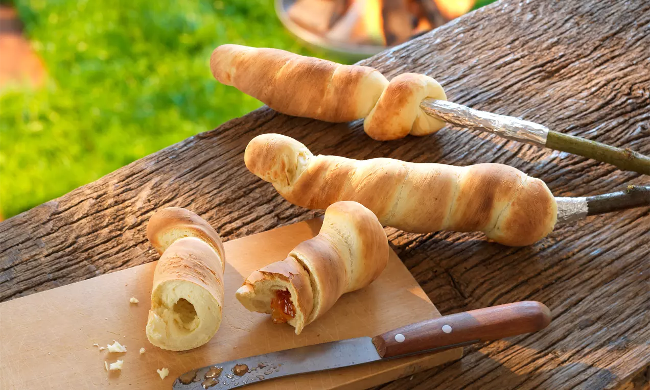 Süßes Stockbrot aus goldbraunem, karamellisiertem Teig auf einem Stab, mit Zimt-Zucker bestreut und warmem Feuer im Hintergrund