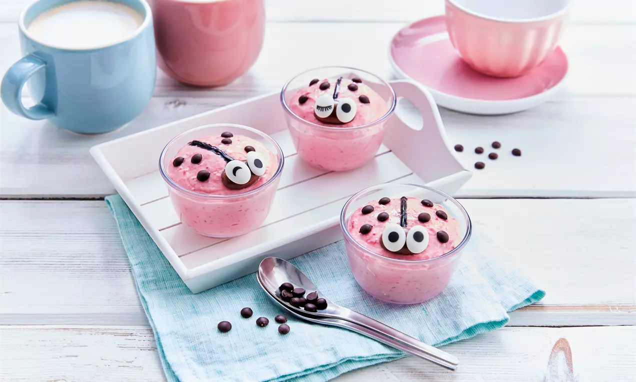 Rosa Dessertcreme in Gläsern mit Schokoladenpunkten und weißen Augen als Marienkäfer dekoriert auf hellem Tablett mit blauer Serviette