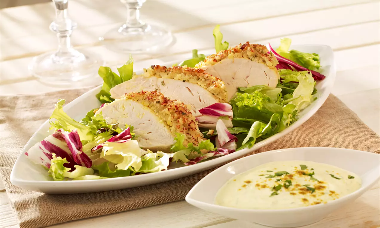 Saftige Hähnchenbrust mit goldbrauner Mandelkruste auf gemischtem Salat, serviert auf weißer Platte neben einer cremigen hellen Soße