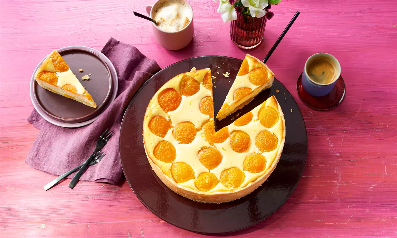 Glutenfreier Käsekuchen mit goldgelben Aprikosen, auf pinker Holzplatte serviert, mit einem angeschnittenen Stück und Kaffee in Tassen