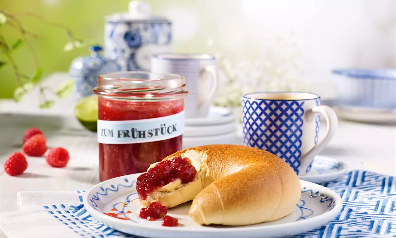 Himbeer-Aprikosen-Konfitüre in einem Glas mit weißem Etikett, daneben ein angerissenes Brötchen mit Konfitüre auf blau gemustertem Geschirr