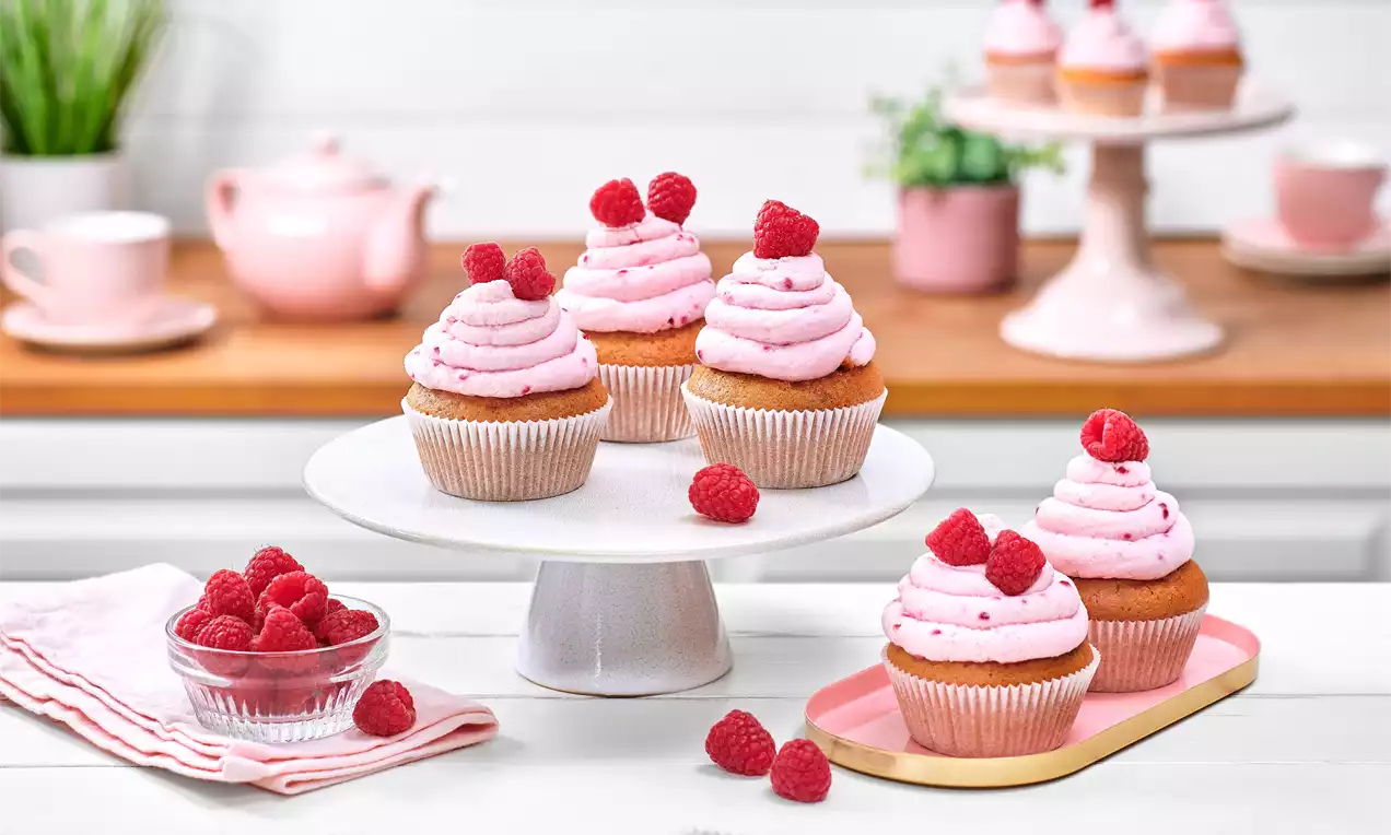 Himbeer-Mascarpone-Muffins mit rosa Cremehaube, frischen Himbeeren und bauchigen Papierförmchen auf weißer Tortenplatte und rosa Tablett
