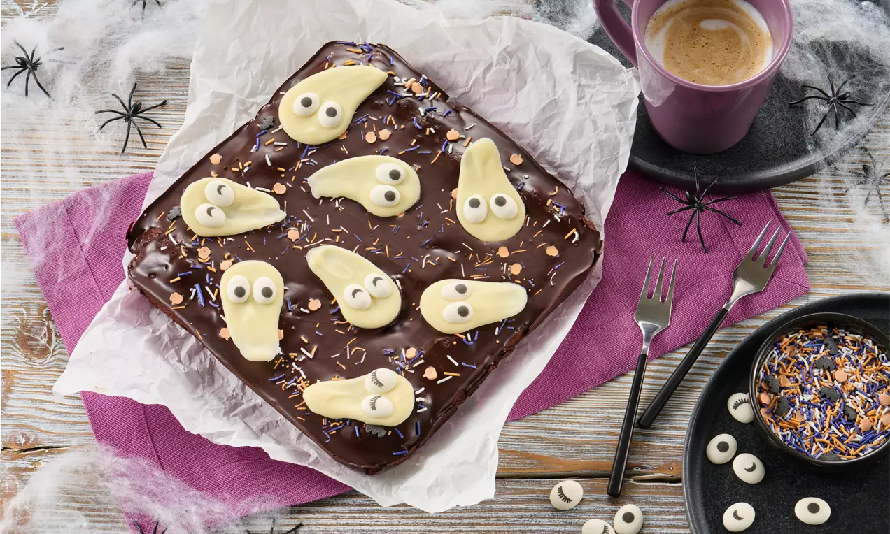 Schokoladiger Brownie mit glänzender Glasur, dekoriert mit weißen Geisterformen, Zuckeraugen und bunten Streuseln auf Backpapier