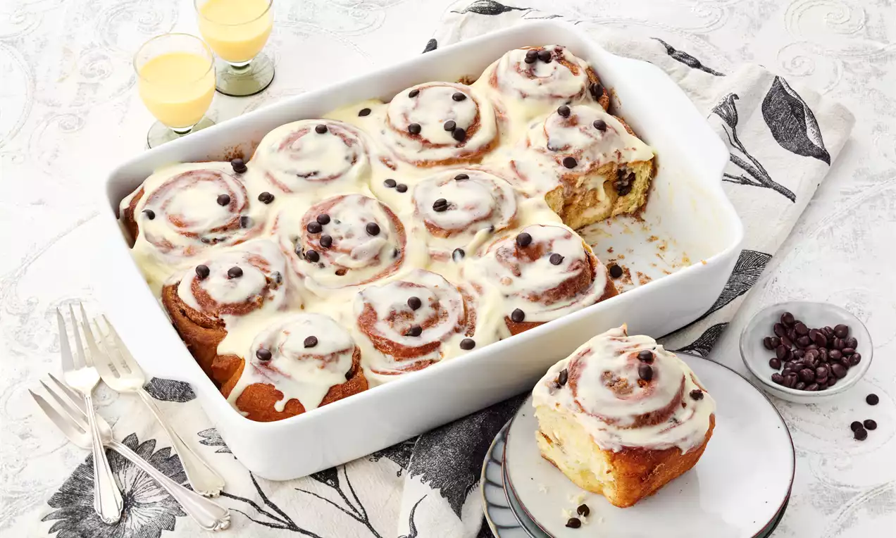 Goldbraune Zimtschnecken mit cremigem Eierlikör-Frosting und Schokotropfen in einer weißen Auflaufform, ein Stück auf einem Teller angerichtet