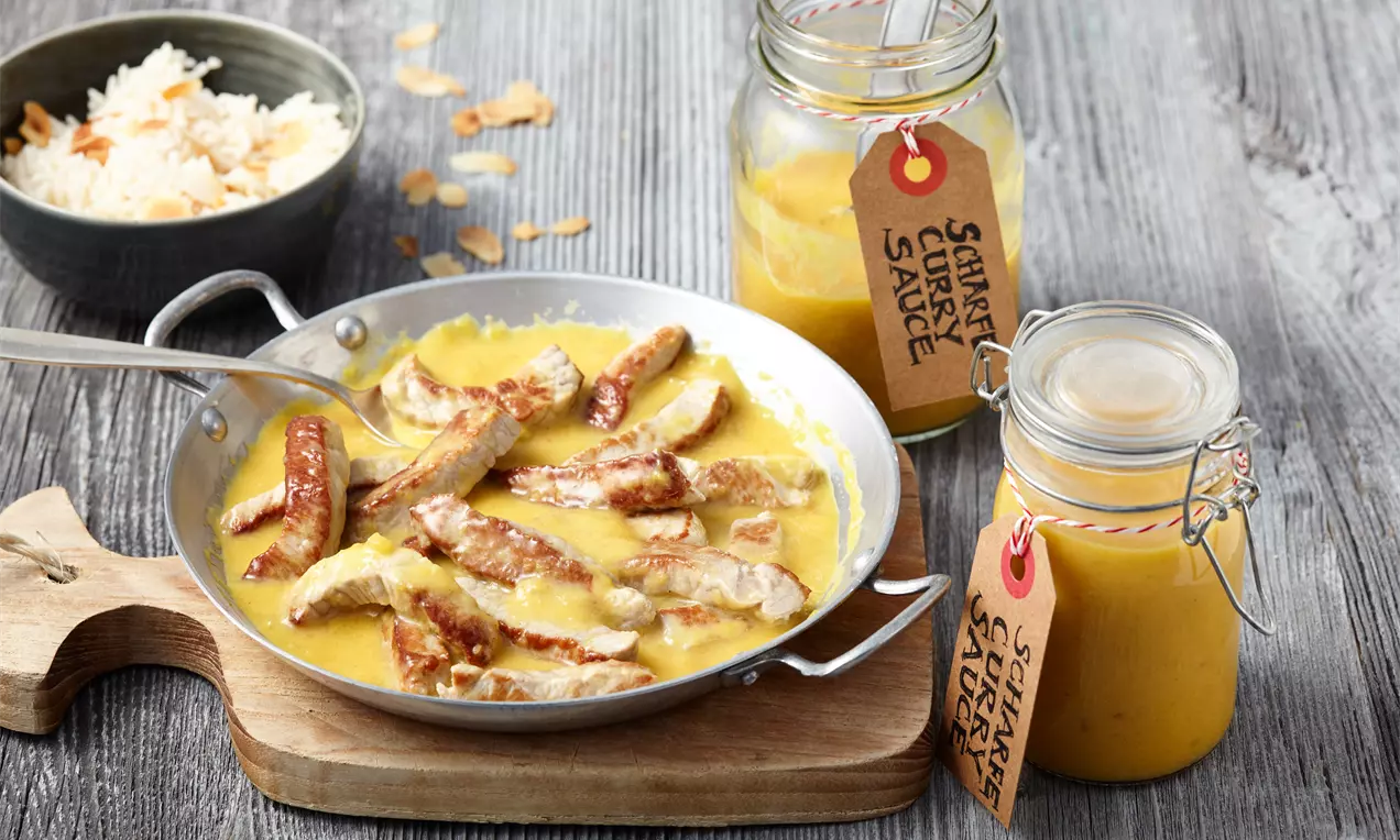 Gebratene Hähnchenstreifen in gelber Currysoße, serviert in einer kleinen Pfanne auf Holzbrett, daneben Gläser mit Currysoße und Reis in Schale