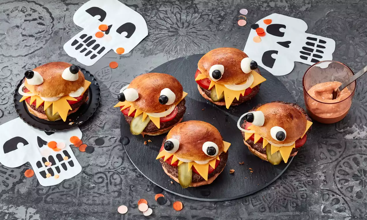 Goldbraune Burger mit dunklen Augen aus Frischkäse und Oliven, Zacken aus Käse und Paprika auf dunklem Teller, dekoriert mit Totenkopf-Motiven