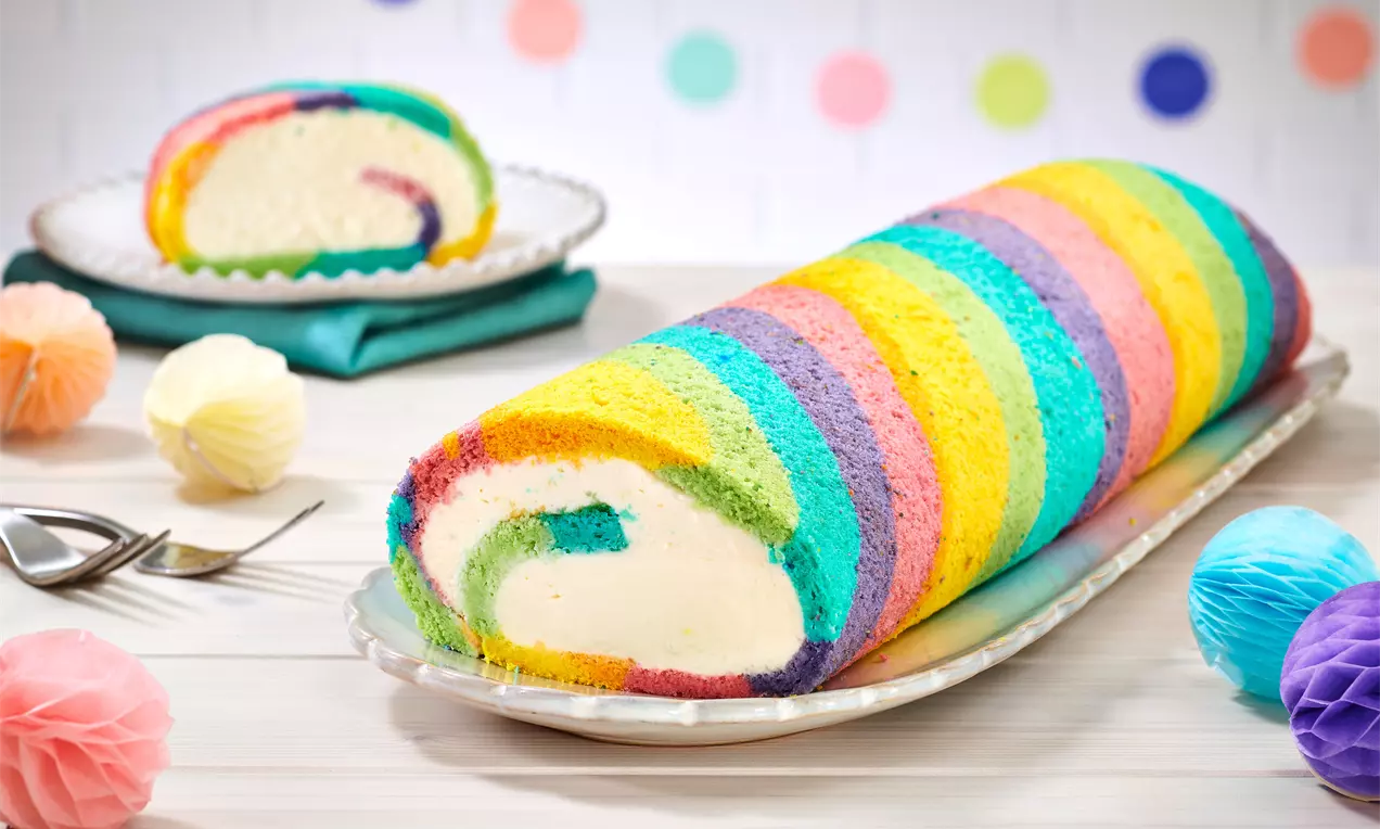 Bunte Regenbogenrolle mit leuchtenden Streifen und heller Cremefüllung, angerichtet auf einer länglichen Platte mit farblichen Dekorationen