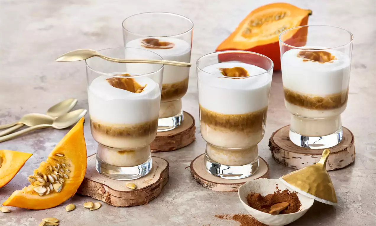 Kleine Gläser mit geschichteter Kürbis-Gewürzmilch und fluffigem Milchschaum, angerichtet auf Holzscheiben mit Kürbisschnitzen und Zimtdeko