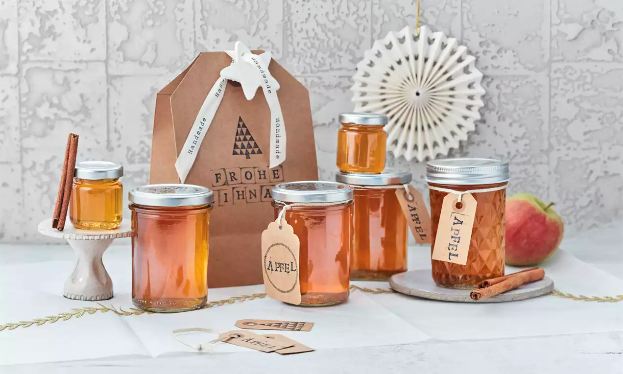 Gläser mit goldgelbem Bratapfelgelee, teils mit Etiketten versehen, auf weißer Oberfläche mit Apfel, Zimtstangen und weihnachtlicher Dekoration