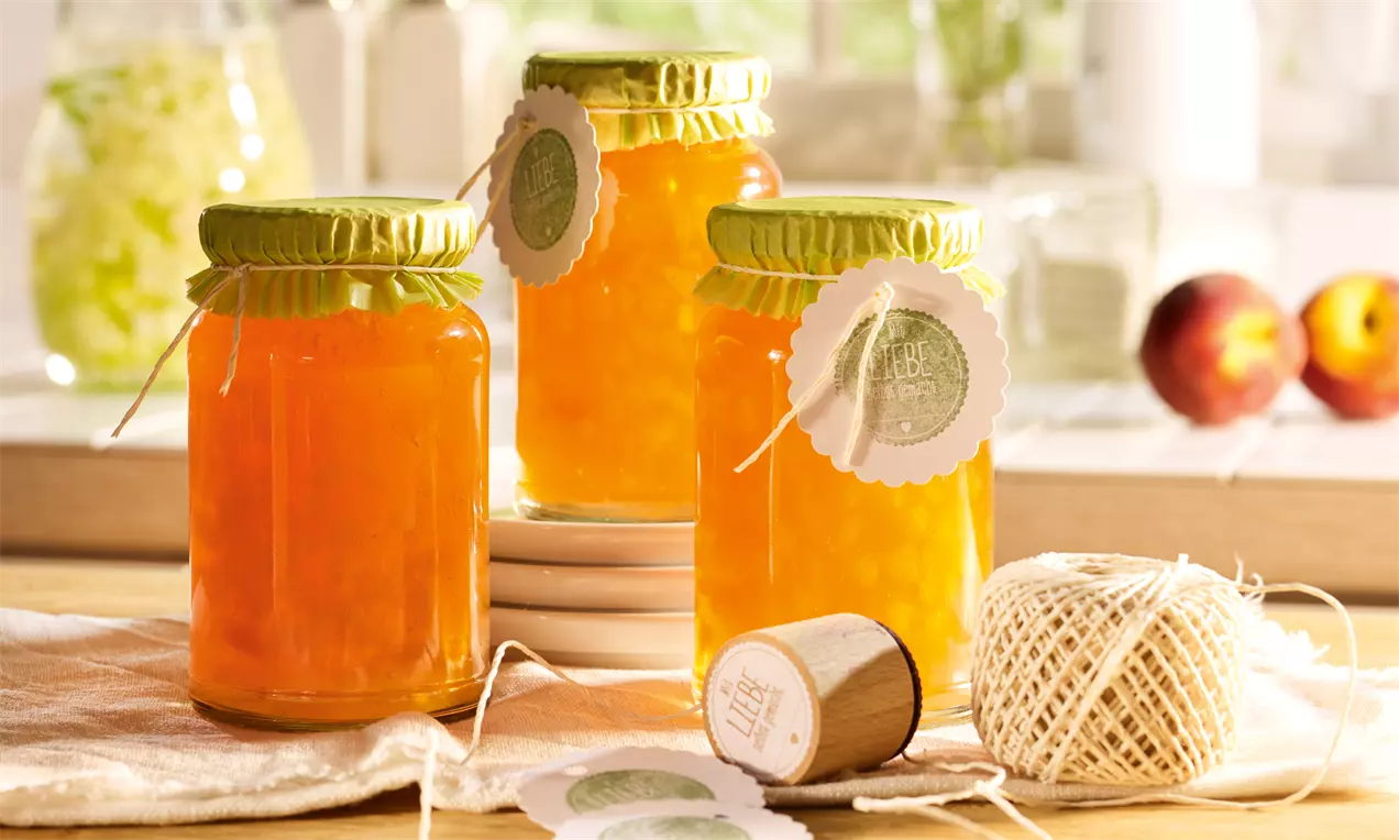 Gläser mit gold-orangem Holunderblüten-Apfel-Gelee und sichtbaren Pfirsichstückchen, mit grünen Deckeln und runden Anhängern verziert