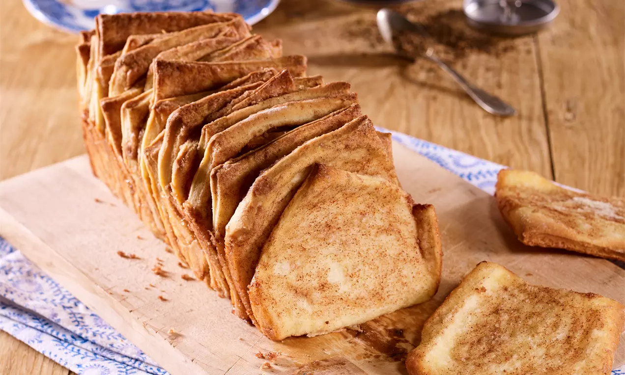 Goldbraunes Pflückbrot mit zart karamellisierten Kanten, in gefächerten Scheiben auf einem Holzbrett, flankiert von einzeln entnommenen Stücken