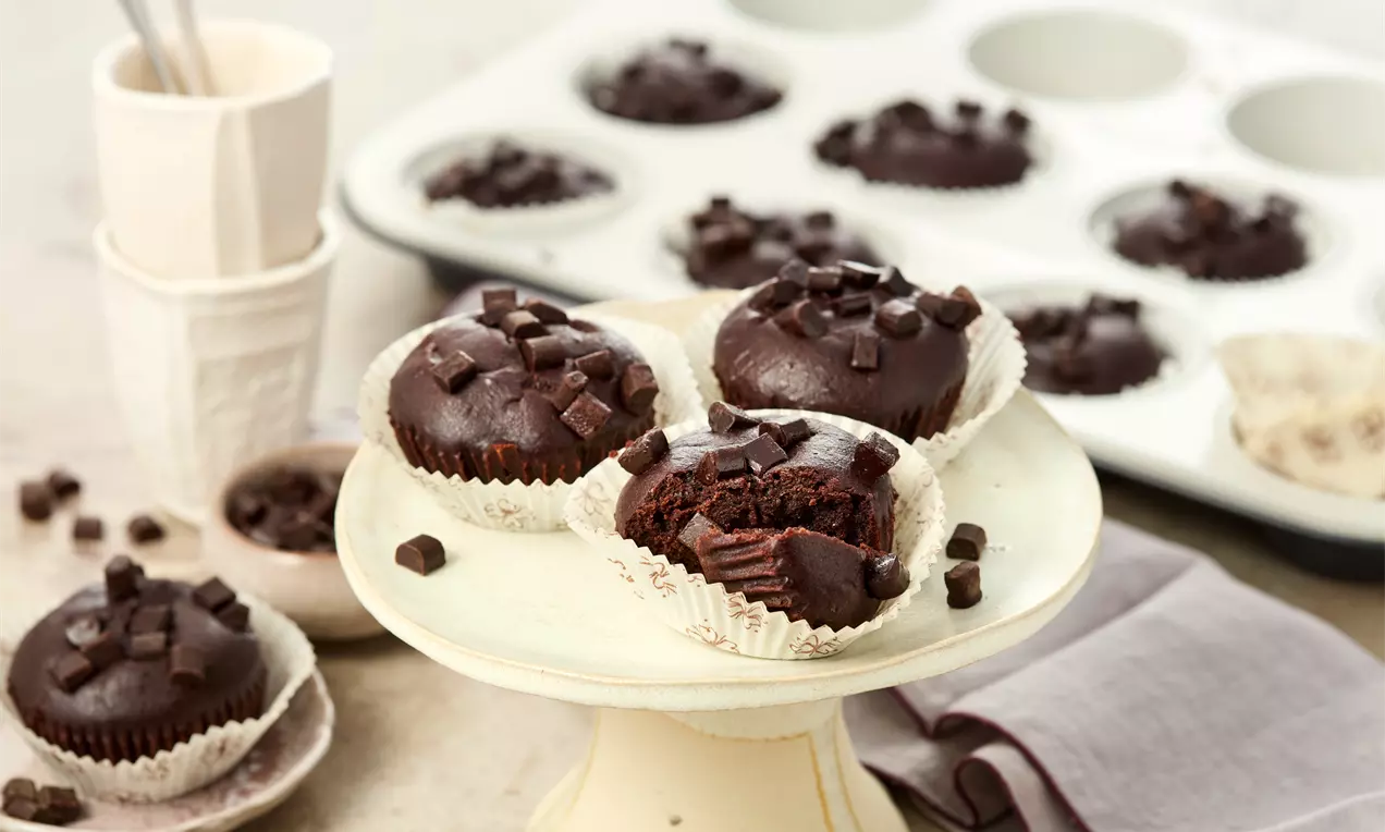 Vegane Schokomuffins auf einer hellen Kuchenplatte, garniert mit Schokostückchen. Ein Muffin ist angebissen. Im Hintergrund Muffinform mit weiteren Muffins und leere Papierförmchen.