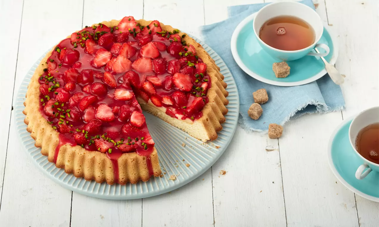Heller Biskuitboden mit glänzend rotem Erdbeerguss und frischen Erdbeerstücken, bestreut mit grünen Pistazien auf einer weißen Platte neben türkiser Teetasse