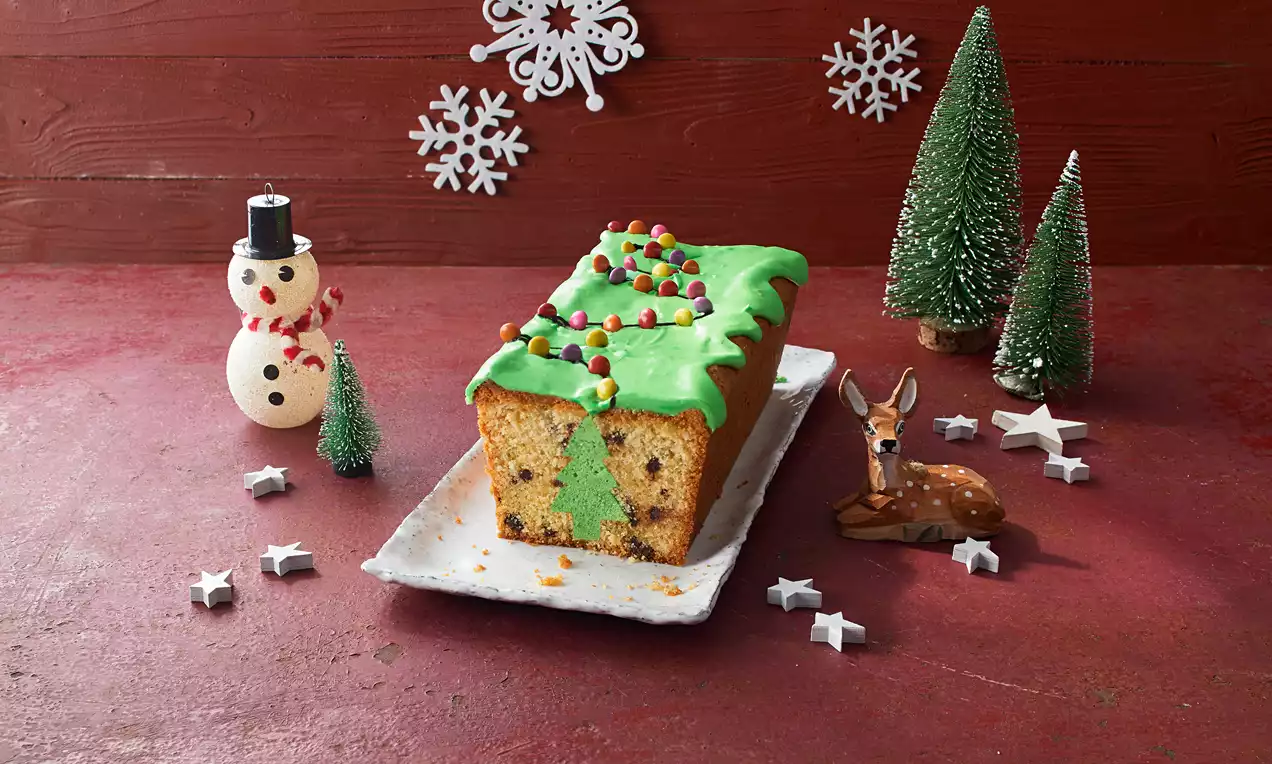 Rechteckiger Kastenkuchen mit grüner Glasur und gezacktem Rand, bunten Zuckerperlen und Tannenbaummotiv auf weißer Porzellanplatte