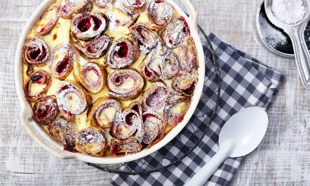 Überbackene goldbraune Pfannkuchenröllchen mit fruchtroter Füllung und weißem Puderzucker in einer runden Form auf karierter Stoffserviette