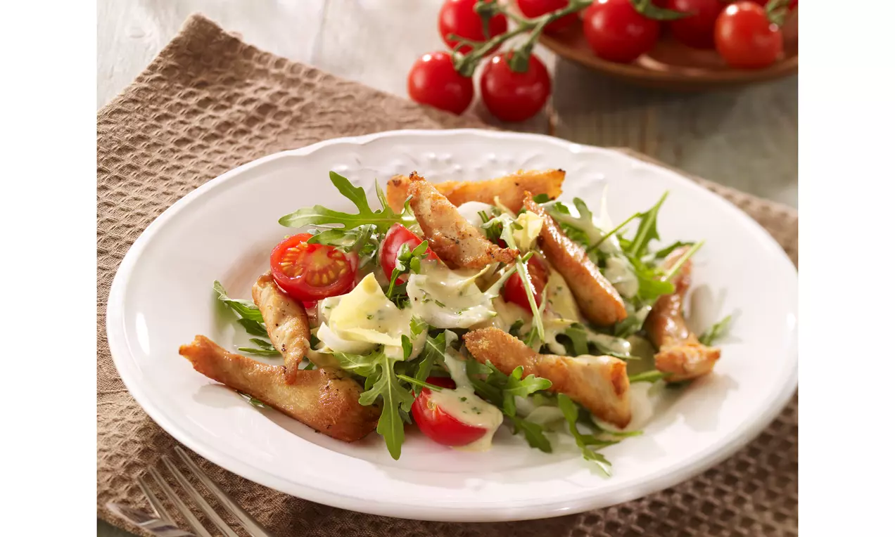 Hähnchenstreifen goldbraun gebraten auf grünem Rucola mit roten halbierten Cherrytomaten und heller Dressingcreme auf einem weißen Teller