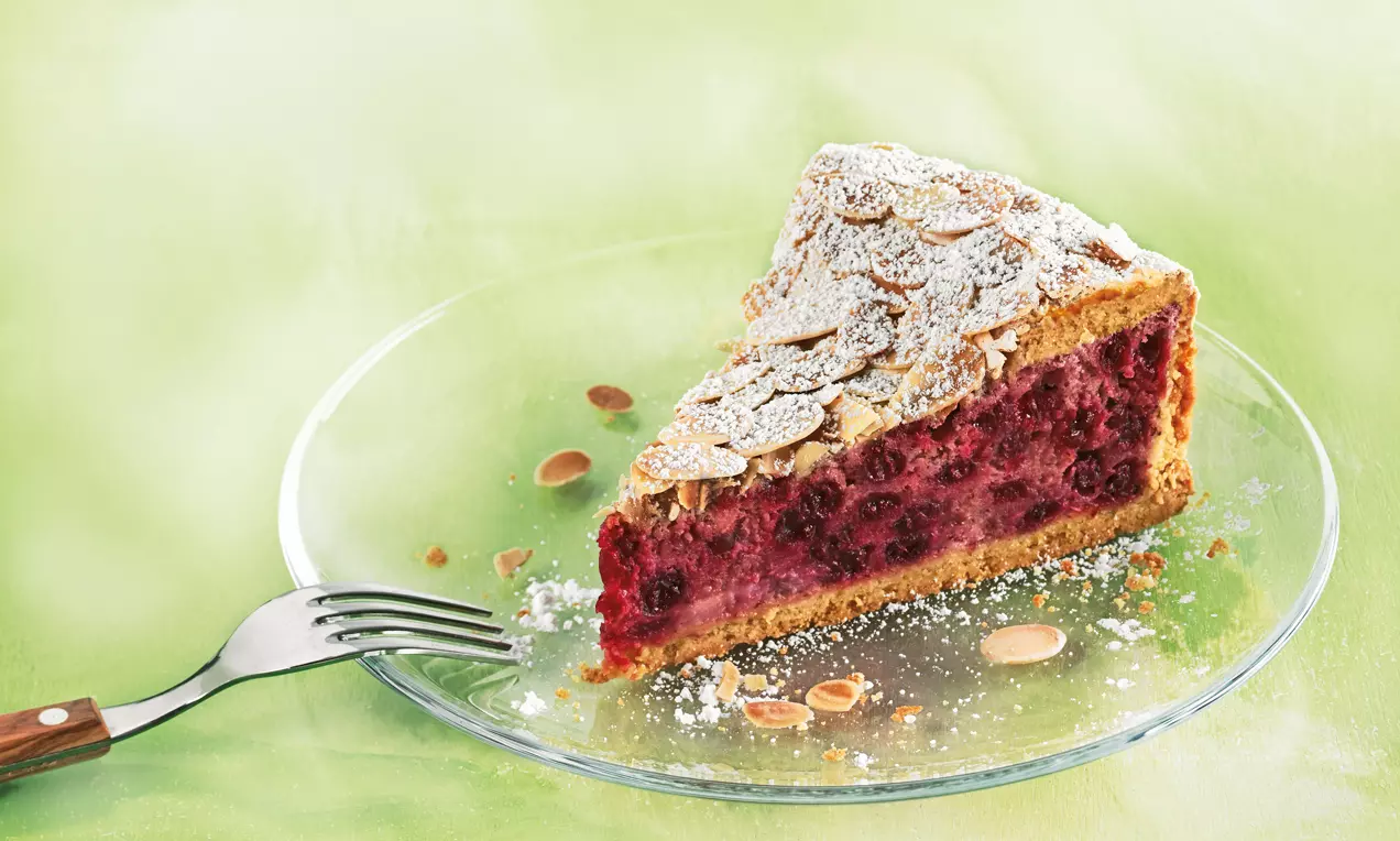 Eine dreieckige Träubleskuchenscheibe mit goldbraunem Mürbeteig, roter Johannisbeerfüllung, Mandelblättchen und Puderzucker auf einem Glasteller