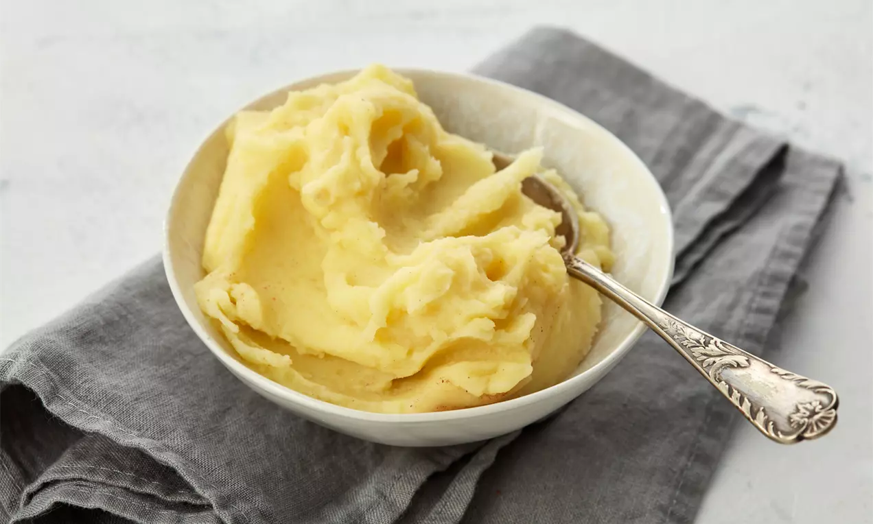 Cremiges, hellgelbes Kartoffelpüree mit glatter und luftiger Textur in einer weißen Schale, angerichtet auf grauem Stoff mit silbernem Löffel