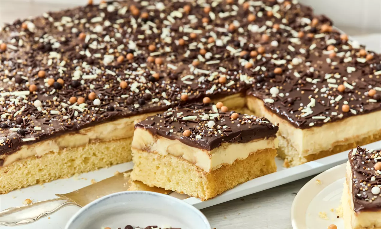 Bananenschnitte mit saftigen Bananenscheiben, cremiger Vanillepudding-Schicht und dunkler Schokoganache auf weißem Teller mit Schokoraspeln