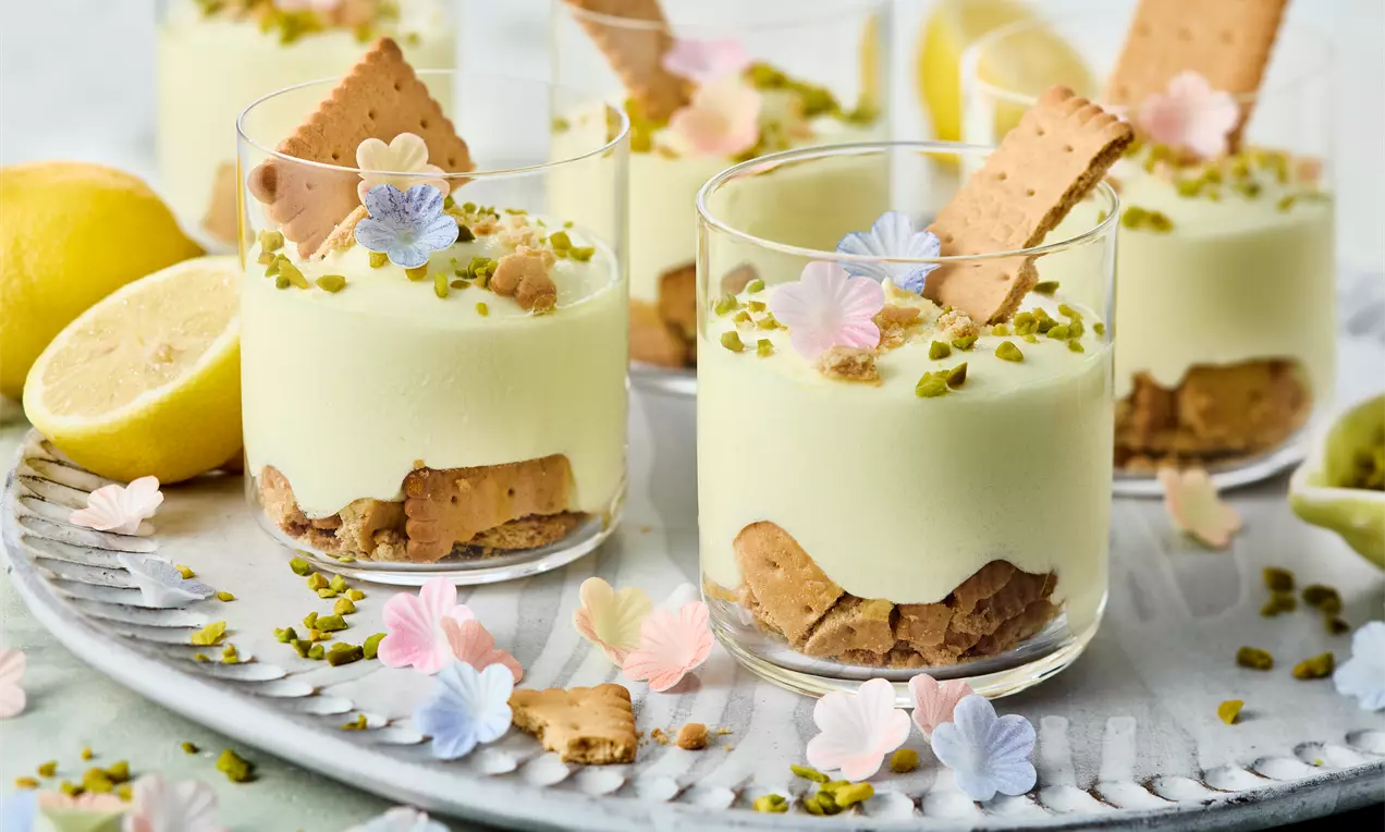 Zitronencreme-Dessert in Gläsern mit knusprigem Keksboden, gehackten Pistazien, Dekoblumen und halbierten Zitronen auf einem Tablett angerichtet