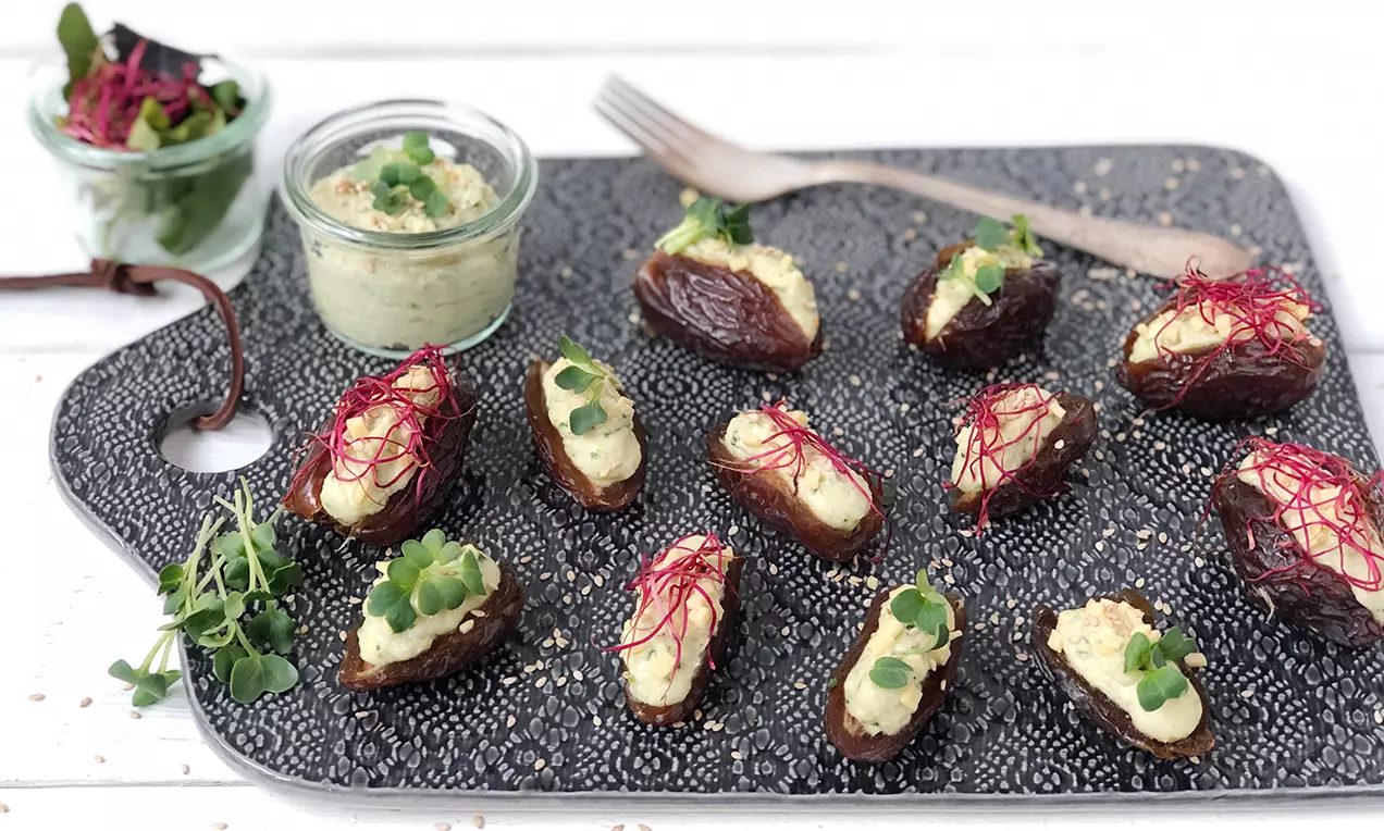 Gefüllte braune Datteln mit hellgrüner Zucchini-Mandelcreme, garniert mit roten Fäden und grünen Blättern auf dunkler Servierplatte