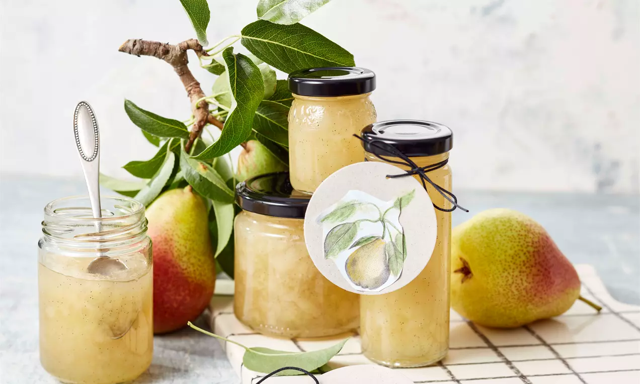 Gläser mit goldgelber Birnenmarmelade, teils geöffnet auf Gitter, dekoriert mit grünen Birnenblättern und frischen rotgelben Birnen